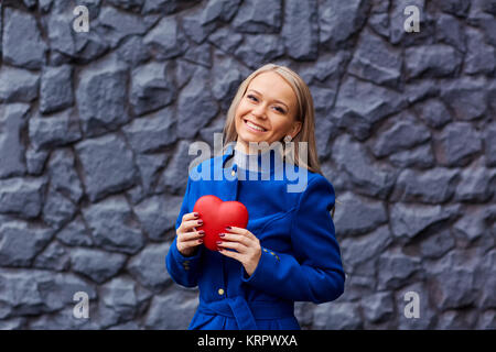 A blond girl in a blue coat holds a red heart in her hands.  Stock Photo