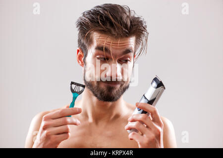 Young man is holding electric and classic razor. He is deciding which one to use to shave his beard. Stock Photo