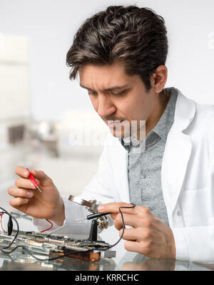Young energetic male tech or engineer repairs electronic equipment in research facility. Shallow DOF, focus on the face of the worker. Stock Photo