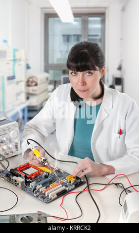 Young female tech soldering a circuit board in modern laboratory looks at the viewer Stock Photo