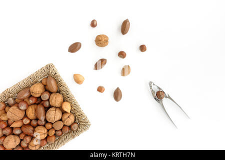 Whole Walnuts, pecan nuts, hazelnuts and almonds with shell in a brown wicker basket, scattered nuts and a nutcracker on white background. Top View. Stock Photo