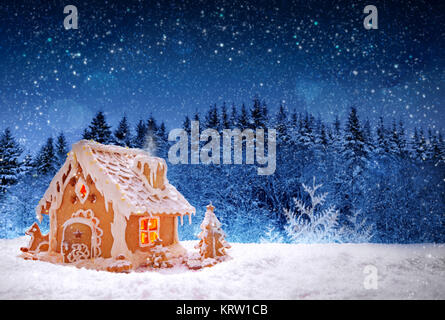 Gingerbread house isolated on fir trees forest background. Christmas card with snowfall and luminous house in the forest. Stock Photo