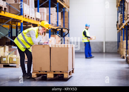 Young woman warehouse worker with smartphone, making a phone call. A man worker holding a box. Stock Photo