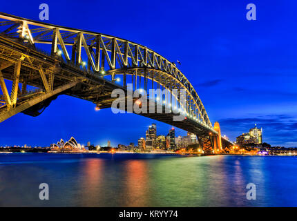 Side view of Sydney Harbour bridge towards city CBD, the Rocks and Circular quay at sunset across blurred reflecting harbour water. Bridge arch is ill Stock Photo
