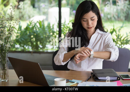 asian businesswoman look at her watch at workplace. startup woman check time on wristwatch at office.  young female entrepreneur with paperwork on tab Stock Photo