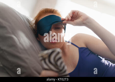 Young woman with sleep mask waking up in bed and snoozing alarm clock on cell phone, mobile telephone. Girl sleeping in bedroom at home and waking up  Stock Photo