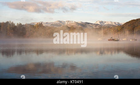 Mist rises around boats moored on Windermere lake, beside woodland trees in autumn colours, at Ambleside in England's Lake District National Park. Stock Photo