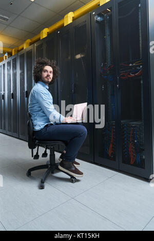 Young technician working on a laptop next to black server racks Stock Photo