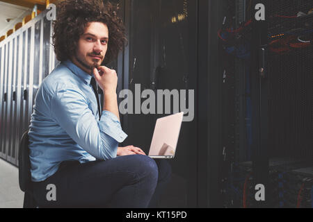Young technician working on a laptop next to black server racks Stock Photo