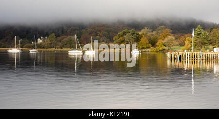 Mist rises from woodland in autumn colour at Ambleside on Windermere lake, under the mountains of England's Lake District National Park. Stock Photo