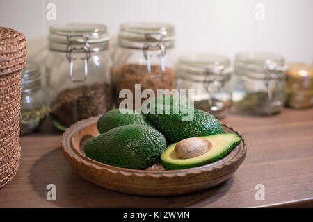 A bowl full of avocados including one cut avocado on a traditional old-fashioned kitchen top with glass jars full of ingredients Stock Photo