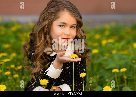 Sunny closeup portrait of pretty little girl with yellow dandeloin. Little girl with many dandelions. Happy girl with beautiful eyes holding one big dandelion. Attractive, adorable girl and dandelion. Beautiful smiling girl with dandelion in hand Stock Photo