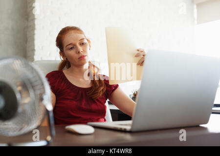 Portrait of young redhead woman working with computer laptop in office at summer during heatwave. The temperature is hot and the hair conditioner is b Stock Photo