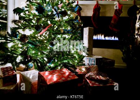 Christmas tree full of ornaments and lights with wrapped presents underneath next to a cozy gas fireplace. Stock Photo