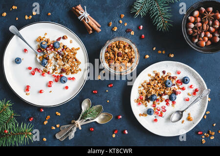 Breakfast composition with homemade cranberry granola, pomegranate, yogurt, spices, nuts and tied tea spoons. Decorated with fir tree branches. Top vi Stock Photo