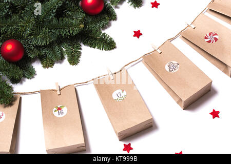 Handmade tinkered advent calendar with paper bags and stickers hanging from fir branches and red stars on a white background from the side Stock Photo