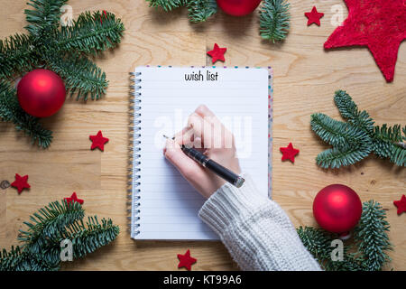 Writing a wish list for Christmas on a notepad with Christmas decorations and fir branches on a wooden table from above Stock Photo