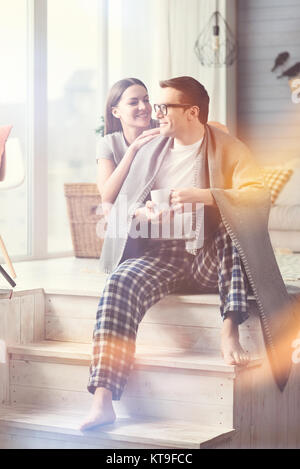 Weekend together. Two charming young people in love sitting by the window and enjoying coffee while his wife bringing him cozy blanket Stock Photo