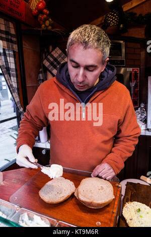 Handsome male seller chef baker in white gloves cooks sandwich with vegetables and cheese standing in medieval shabby kitchen on fair market in Meisse Stock Photo