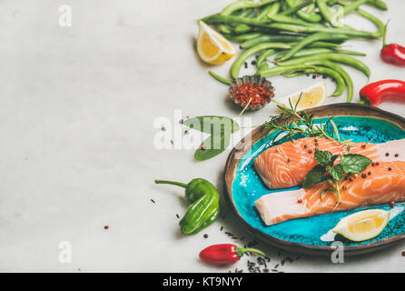 Raw salmon fish fillet steaks with vegetables, greens, rice, spices and lemon in blue plate over light grey marble background, copy space. Clean eatin Stock Photo