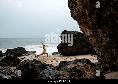 Woman in rain coat doing stand yoga asana on ocean beach during storm. Concept of yoga travelling on Bali Stock Photo
