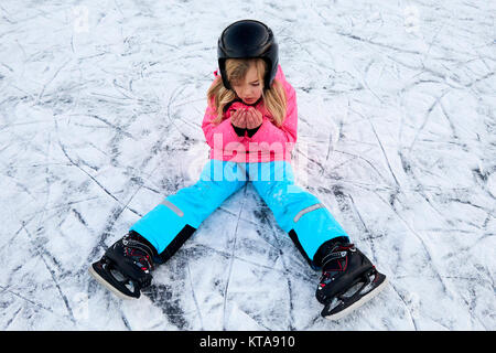 Child girl falling down on ice in snowy park during winter holidays. Wearing safety helmet. Winter children activities. Stock Photo