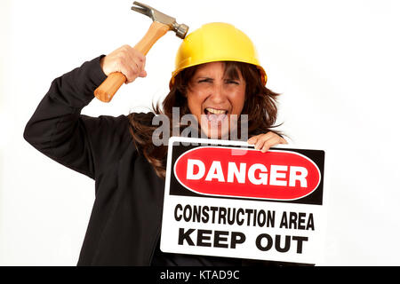 Middle aged woman with long hair wearing a yellow hard hat holding a danger construction area keep out sign hitting the hard hat with a hammer isolate Stock Photo