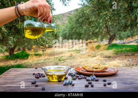 Bottle pouring cretan extra virgin olive oil in a bowl on wooden table at an olive tree field, Greece. Stock Photo