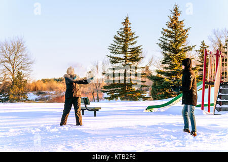 In the foreground, a young woman in glasses in a black overalls joyfully and cheerfully throws a snowball at a young man in a black jacket and jeans.  Stock Photo