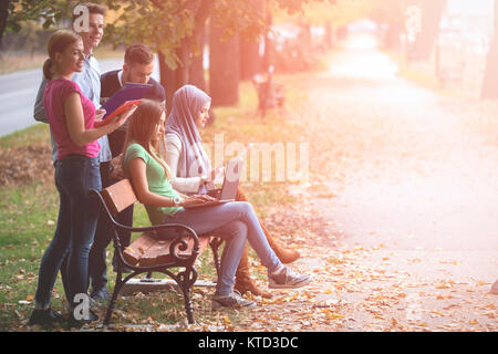 Group of young people using laptop and tablet on a park bench, having fun, talking, hanging out Stock Photo