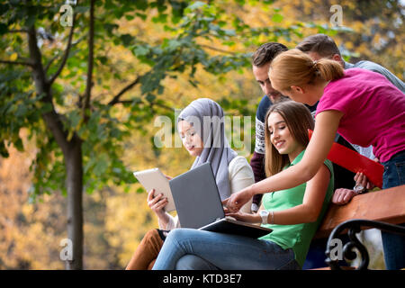 Group of young people using laptop and tablet on a park bench, having fun, talking, hanging out Stock Photo