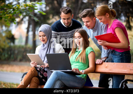 Group of young people using laptop and tablet on a park bench, having fun, talking, hanging out Stock Photo