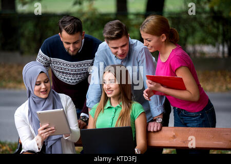 Group of young people using laptop and tablet on a park bench, having fun, talking, hanging out Stock Photo