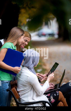 Group of young people using laptop and tablet on a park bench, having fun, talking, hanging out Stock Photo