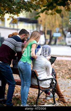 Group of young people using laptop and tablet on a park bench, having fun, talking, hanging out Stock Photo