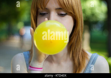 White girl inflating a yellow balloon in a city park. She has orange hair and she is young. Stock Photo