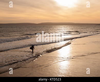 A surfer in silhouette carrying a surfboard walking out of the sea Stock Photo