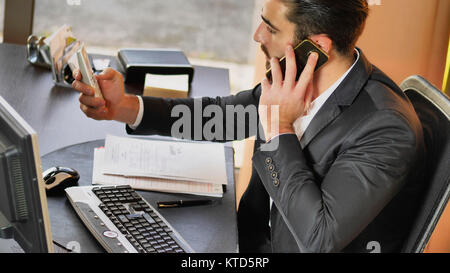 Too much work. Young man using two mobile phones and one desktop computer sitting at office desk Stock Photo