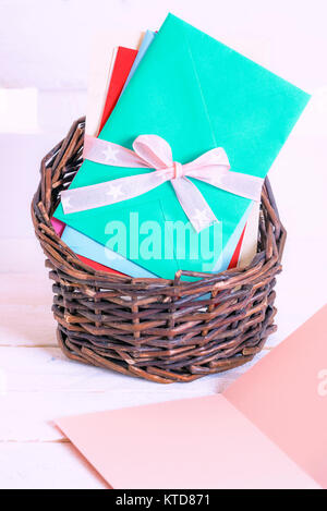 Unwritten message card and a wicker basket full of colorful envelopes, tied together with pink ribbon and bow, on a white wooden background. Stock Photo