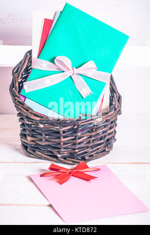 Wicker basket full of multicolored letters tied together with ribbon and a blank message card with a red bow, on a white wooden table. Stock Photo