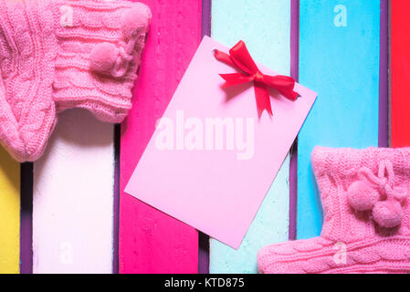 Unwritten pink paper note with red bow surrounded by handmade knitted pink bootees, on a multicolored wooden background. Stock Photo