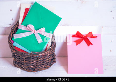 Pile of multicolor envelopes tied with ribbon, displayed in a wicker basket and a paper note with a red bow near it, on a white wooden background. Stock Photo