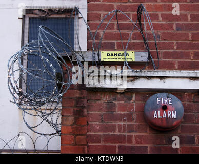 Razor wire against a white wall, with a rusty red fire alarm and yellow warning sign on a red brick wall, and a window with security mesh