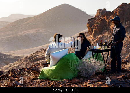 Couple enjoying sundowner while overlooking vast red rock landscape of Damaraland - Huab Under Canvas, Damaraland, Namibia, Africa Stock Photo