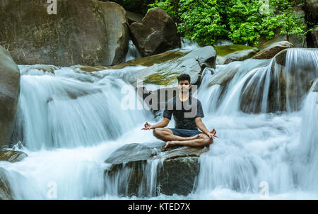 Young adult boy sitting on a rock midst smooth splashing and flowing water, meditating at a portion of Nagarmadi Falls in Chendia, Karnataka, India Stock Photo