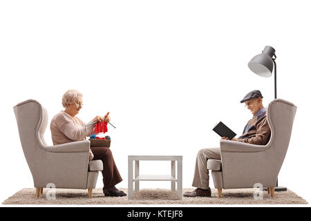 Seniors seated in armchairs with one of them knitting and the other reading a book isolated on white background Stock Photo