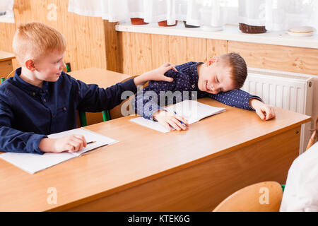 Tired and sleepy boy during the lesson of primary school. His friend wake up shoolmate Stock Photo