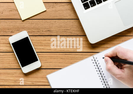Overhead view of a neat wooden desktop with male hand writing on a notebook, smartphone, laptop, and notepad. Stock Photo