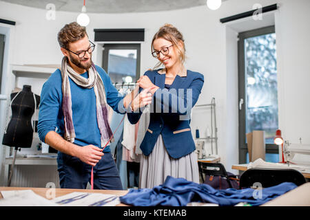 Handsome tailor measuring jacket on the woman client standing at the studio full of tailoring tools and equipment Stock Photo