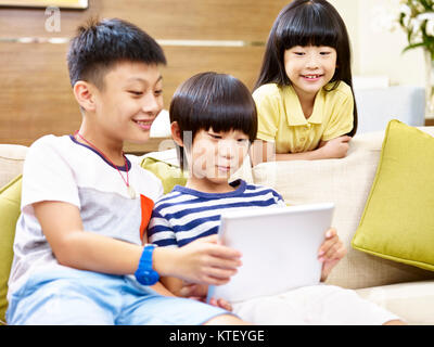 three asian children two little boy and one little girl sitting on couch at home playing video game with digital tablet, focus on the little girl in t Stock Photo